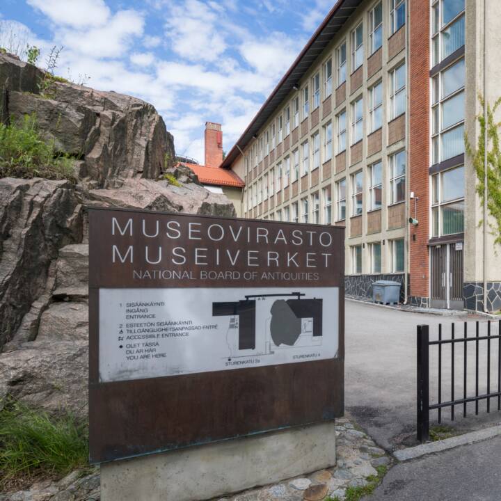 Kesäisessä maisemassa virastorakennus, jonka edessä kalliota ja kyltti. Kyltissä lukee suomeksi Museovirasto, ruotsiksi Museiverket ja englanniksi pienemmällä kirjasimella National Board of Antiquities.